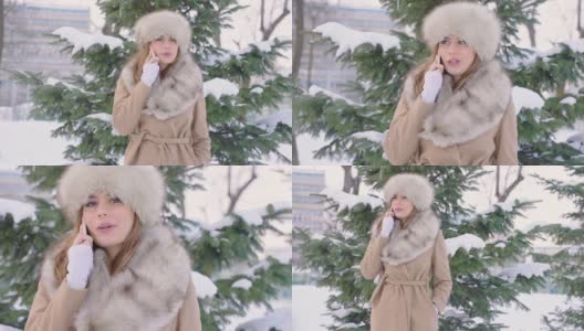 美丽的女人戴着皮帽看相机和使用智能手机在冬天的公园在城市在下雪的天气与飘落的雪，慢镜头高清在线视频素材下载