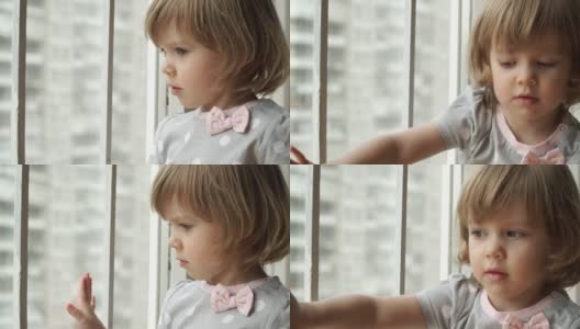 严肃的小女婴坐在窗台上，望着大城市的窗外，手梦幻般地滑在玻璃上。沉思的孩子注视着窗外的街道。高清在线视频素材下载