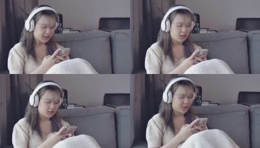 一个美丽的亚洲少女听音乐耳机和智能手机在她的客厅在公寓房间。高清在线视频素材下载
