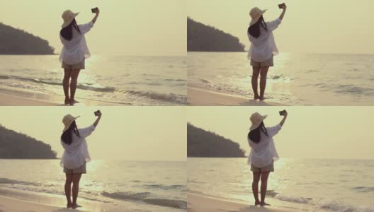 亚洲美丽的年轻女子使用智能手机自拍，而站在海滩日落。生活方式旅游度假夏季概念。高清在线视频素材下载