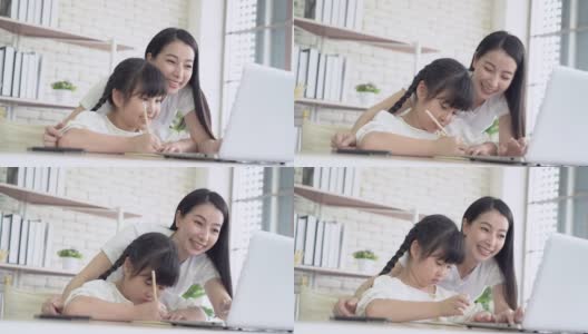 在家教育。快乐的年轻亚洲妈妈和女儿在妈妈的帮助下，坐在家里的书桌上，用笔记本电脑做作业。女孩高兴地在家学习。在线教育的概念高清在线视频素材下载