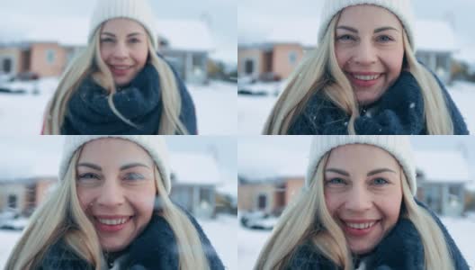 一个年轻的金发女人的肖像在红色大衣与围巾微笑进入相机，而站在飘落的雪。在后院田园诗般的房子与冬天的景色。高清在线视频素材下载