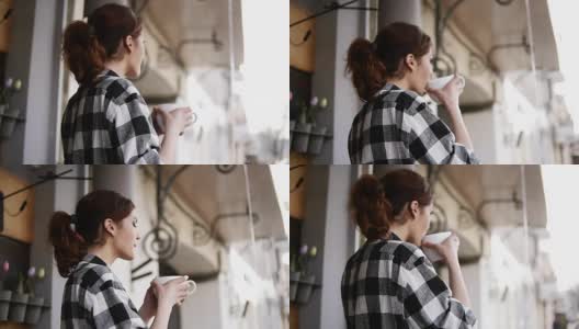 一个穿着格子衬衫的可爱女孩站在窗户对面，用一只白色的大杯子喝着一杯普通咖啡，看着窗外。回来。和画面。模糊的角度来看高清在线视频素材下载