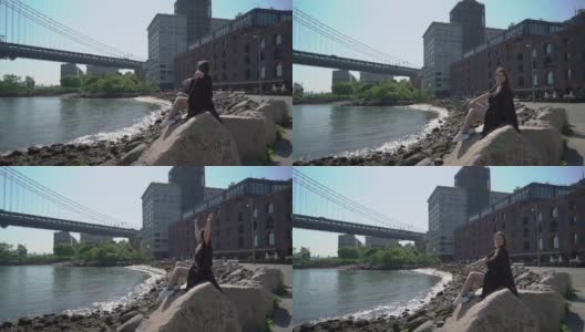 纽约，一名身穿黑衣的女子坐在水边的石头上。伸展手臂了高清在线视频素材下载