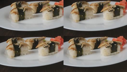 一个白色的大盘子，上面放着四个巨大的寿司，里面有鳗鱼和一片海苔卷在一起，旁边是粉红色的生姜和绿色的芥末高清在线视频素材下载
