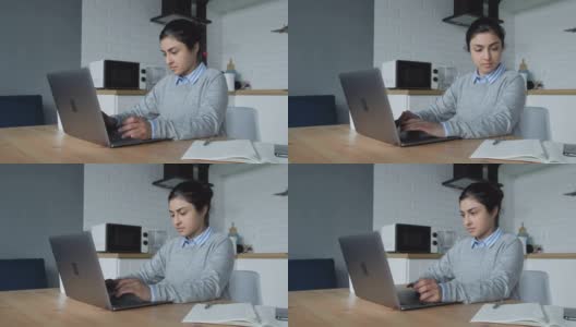 一位年轻的印度妇女在家工作，在电脑上做事，在笔记本上写笔记。她穿着一件衬衫和一件毛衣高清在线视频素材下载