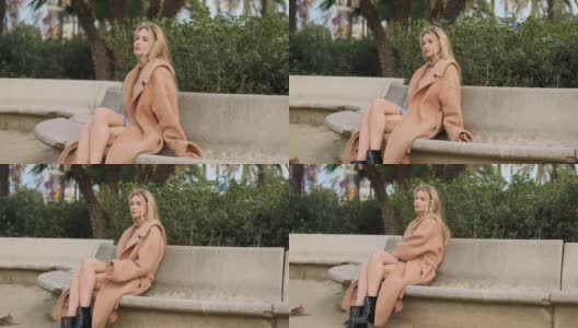 美丽时尚的金发女孩在大衣休息在城市公园的石凳上高清在线视频素材下载