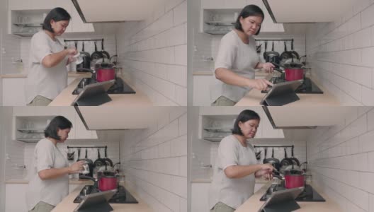 亚洲妇女在厨房做饭，同时看平板电脑的食谱高清在线视频素材下载