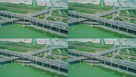 在阳光明媚的日子里，鸟瞰高速公路立交桥的多层交叉路口，快速行驶的汽车被绿树环绕，河边有一条河。高清在线视频素材下载