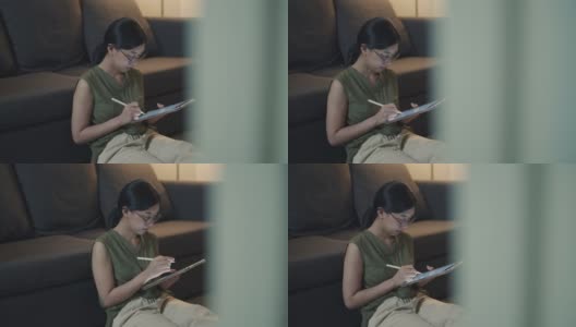 亚洲女性晚上坐在客厅的沙发上用数码平板电脑看新闻。高清在线视频素材下载