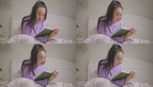 20多岁亚洲女性独自坐在床上看书。穿着紫色睡衣的女性喜欢坐在床上看书。高清在线视频素材下载