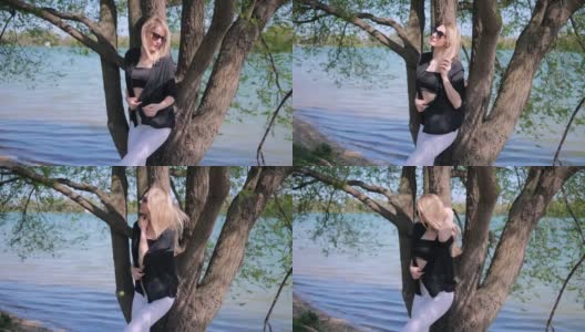 一个美丽的金发女人穿着短上衣，戴着太阳镜，在自然的湖边的一棵树旁摆出性感的姿势高清在线视频素材下载