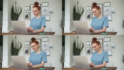 一个年轻的红发女人面带微笑，在家庭办公室的桌子上用笔记本电脑打字。高清在线视频素材下载