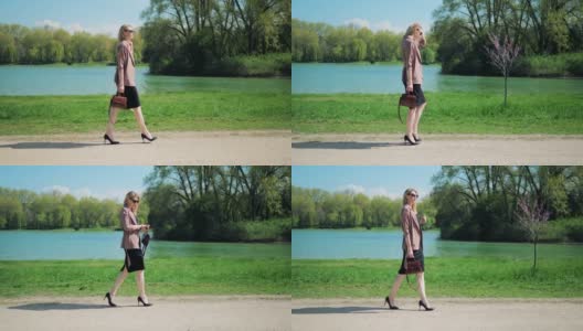 一个年轻的女商人拿着手提包沿着湖走过公园，看着她怀里的手表。高清在线视频素材下载
