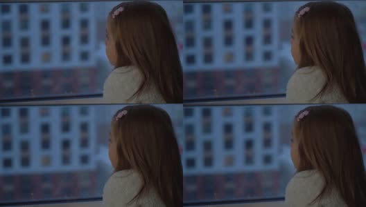 冬夜小女孩站在窗台的背影，望着窗外的雪花和城市风光。近距离高清在线视频素材下载