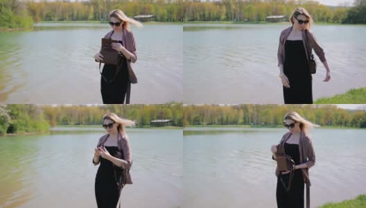 一个美丽的年轻女子在湖边的公园里，为了让自己更漂亮，她正在手提包里找一面镜子高清在线视频素材下载