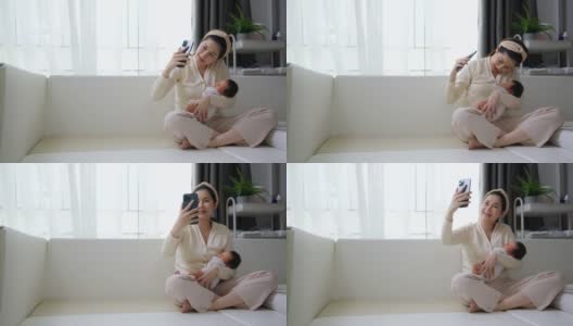 美丽的妈妈抱着可爱的新生儿用手机视频通话。技术高清在线视频素材下载
