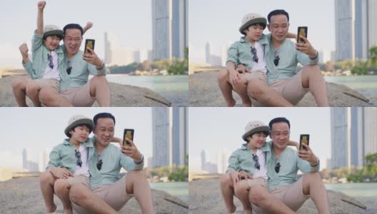 一位亚洲父亲和他快乐的儿子用智能手机自拍，一起在城市附近的岩石岸边度过美好时光高清在线视频素材下载