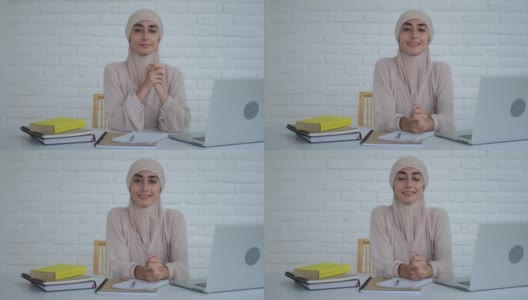 一位年轻的穆斯林妇女戴着希贾布坐在桌旁微笑，她很高兴，因为她可以接受教育。桌上有笔记本电脑和书。穆斯林妇女远程教育的概念高清在线视频素材下载