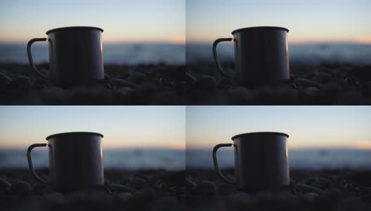一个旅行不锈钢杯站在岩石上的特写，在海滩的背景下，平静的日落在海洋海浪上。一个概念的世界美，自然和户外旅行。有选择性的重点高清在线视频素材下载