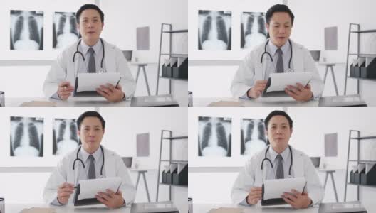 年轻的亚洲男医生穿着白色医疗制服，带着听诊器用电脑笔记本电脑与病人进行视频通话，看着医院的摄像头。高清在线视频素材下载