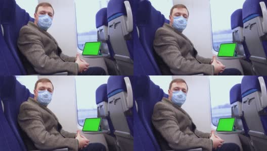 穿着外套、戴着医用口罩的千禧一代男子坐在火车上，看着摄像机，他面前的折叠桌上放着一台带有色度键的绿色屏幕的平板电脑高清在线视频素材下载