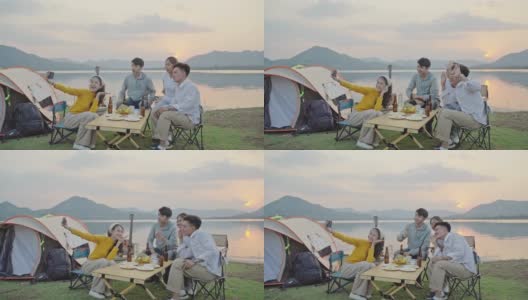小组四人朋友亚洲男人和女人野营野餐庆祝，通过智能手机自拍享受乐趣和地面帐篷。日落假期期间的水库区域。高清在线视频素材下载