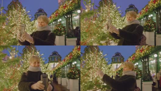 有魅力的成熟女子，一名游客，在圣诞节期间用智能手机在灯火通明的公园里拍照和自拍，享受大城市的节日装饰。高清在线视频素材下载