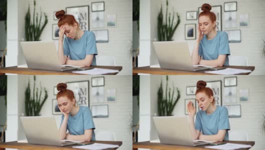 一个年轻的红头发女人，疲倦地坐在书桌前用现代笔记本电脑工作。高清在线视频素材下载