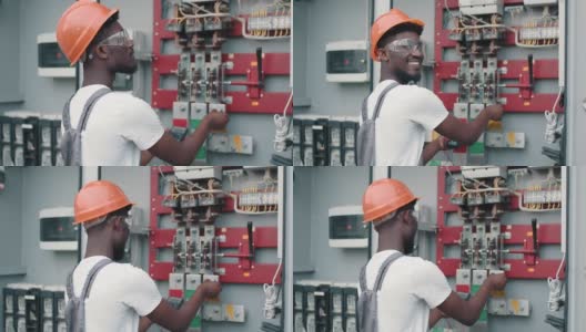 非裔美国人，身穿灰色工作服，戴着安全帽和眼镜，微笑着看着镜头，站在太阳能电站的开关柜旁。替代能源的生产高清在线视频素材下载
