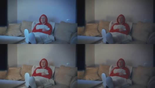 穿着红色连帽运动衫的男人，晚上坐在沙发上把脚放在面前看电视，电视灯高清在线视频素材下载