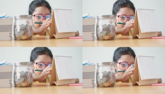 亚洲小女孩用桌子上的玻璃罐在日历上写字，隐喻省钱的概念高清在线视频素材下载