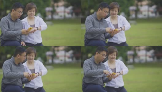 亚洲华人中成年夫妇在公园长凳上锻炼后，使用健身追踪器移动应用连接到健身追踪器高清在线视频素材下载