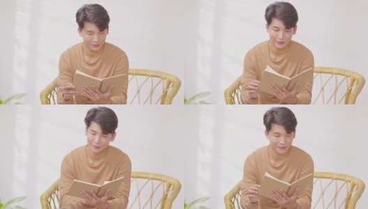 快乐英俊的亚洲男人坐在沙发上微笑和阅读书籍放松时间在家里。兴高采烈的商人穿着蓝色衬衫，坐在沙发上看书高清在线视频素材下载