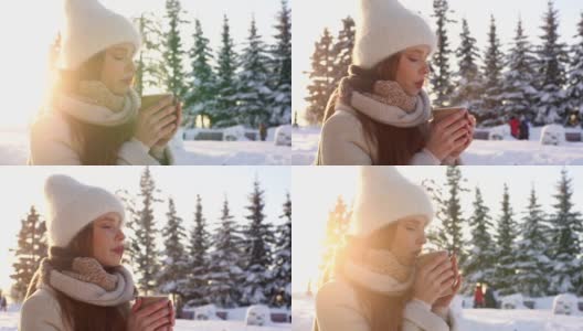 在雪城公园里，迷人的年轻女子戴着白色帽子，闭着眼睛享受着从杯子里拿出来的热的芳香咖啡或巧克力。高清在线视频素材下载