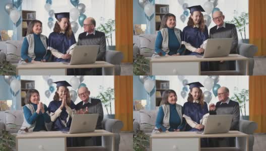 在线毕业典礼，快乐的父母和女毕业生高兴地鼓掌在毕业典礼上通过视频连接，而坐在家里的沙发上，背景是气球高清在线视频素材下载