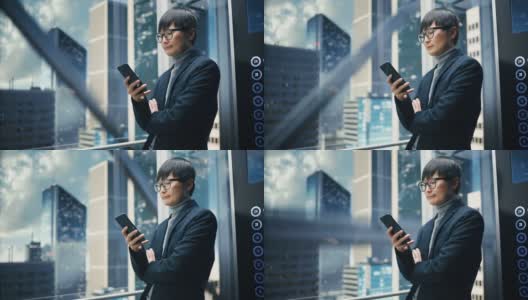 成功时尚的日本商人乘坐玻璃电梯到现代商务中心的办公室。帅哥在使用智能手机时微笑，写短信，在电梯里查看社交媒体。高清在线视频素材下载
