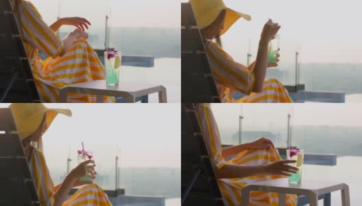 亚洲女性游客在夏天穿着黄色的连衣裙，戴着草黄色的帽子，放松在一个无边的游泳池，享受蓝色的鸡尾酒在日落。高清在线视频素材下载