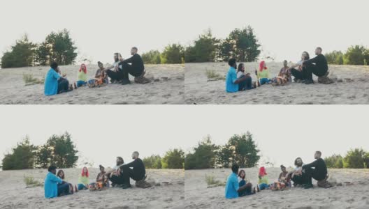 朋友们坐在湖边的沙滩上，一起消磨时间，在篝火旁玩耍，放松，一个女人弹吉他，唱歌，喝啤酒高清在线视频素材下载