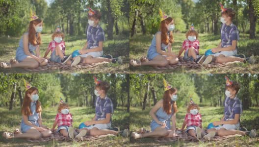 宽镜头快乐的白人兄弟姐妹坐在春天夏天的公园野餐聊天。戴着派对帽和冠状病毒口罩的红发男孩女孩在阳光下户外庆祝生日。高清在线视频素材下载