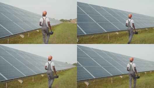 能源工程师带着仪器在太阳能农场上行走。非洲裔美国人在制服和头盔使用仪器，而工作的领域与太阳能电池板的后视图。生产能源高清在线视频素材下载