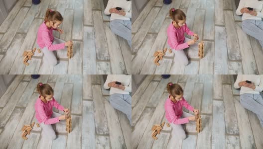 无所谓的母亲用手机，女儿在家里的地板上玩木结构高清在线视频素材下载