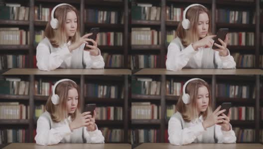 可爱的白人小女孩拿着智能手机玩手机应用，坐在图书馆里玩游戏。孩子在手机里学习，看视频，玩手机技术的概念高清在线视频素材下载