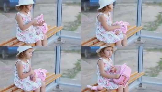 一个带着帽子和背包的小女孩正坐在公交车站的长椅上，等待进一步旅行的交通工具高清在线视频素材下载