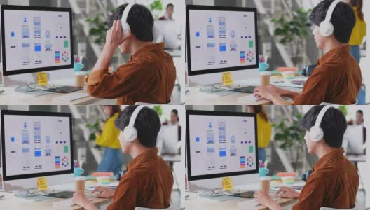 在现代创意办公室，年轻的亚洲程序员男子戴着耳机在电脑桌面上编写移动应用程序，忙碌的亚洲ux, ui设计开发团队，放松的亚洲男性听着音乐工作高清在线视频素材下载