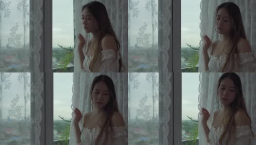 4K沮丧年轻美丽的亚洲女人独自住在公寓的窗户旁。孤独悲伤的女孩站在一边，看着窗外的城市，思考着生活中的问题。负面情绪的概念。高清在线视频素材下载