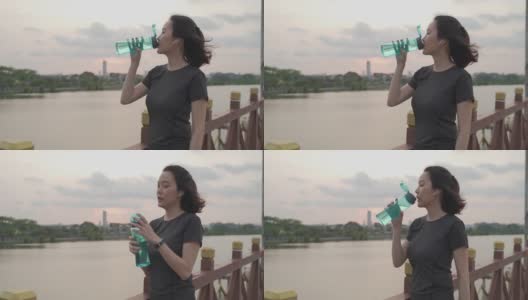 亚洲运动员女子在绿色的自然，树木公园和湖在傍晚日落感觉口渴喝水从瓶子。能量饮料补充疲劳和疲惫的水分。锻炼和喝水。高清在线视频素材下载