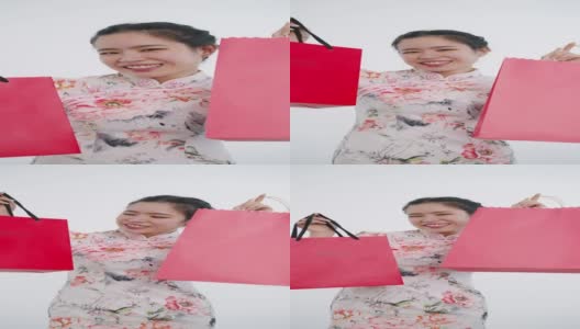 中国/亚洲妇女在中国新年期间，双手拿着红色和粉红色的购物袋，白色背景，穿着旗袍白色花卉图案连衣裙高清在线视频素材下载