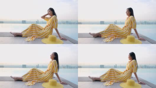 一名年轻的中国女子穿着黄色连衣裙，戴着帽子，坐在摩天大楼的无边泳池边上。女人享受城市的风景，日落时的自然，酒店的屋顶高清在线视频素材下载