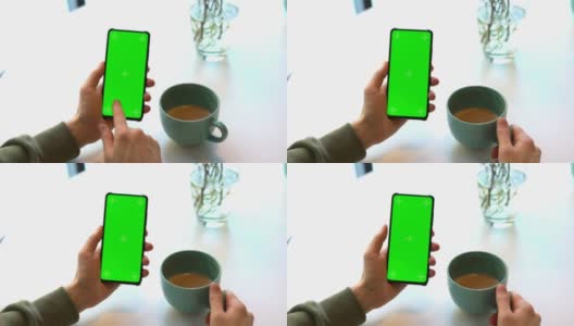 男人拿着带Chroma屏幕的智能手机，在咖啡馆的白色桌子上喝着卡布奇诺咖啡，通过互联网，滑动屏幕，使用网站，绿色屏幕的商家选择性聚焦高清在线视频素材下载